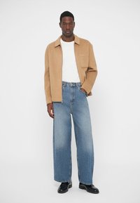 Beige wool blend jacket with a pocket, white textured shirt, loose-fit light blue denim jeans, and black shoes on a plain background.