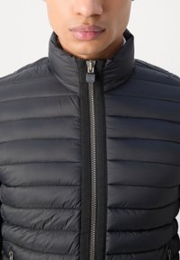 Doudoune noire matelassée avec un col montant, fermeture éclair à l'avant et accents élastiques. Tissu léger avec une texture lisse et un design élégant.