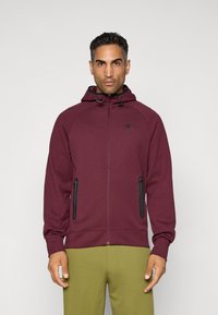 Mann, der nach vorne schaut, trägt einen bordeauxfarbenen Reißverschluss-Hoodie mit kleinem Logo und grüne Hosen vor einem schlichten hellgrauen Hintergrund.
