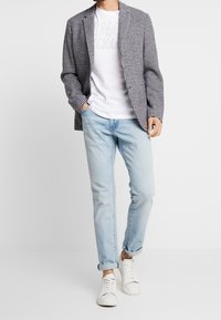 Sako s černobílým kostkovaným vzorem houndstooth, bílé tričko s kulatým výstřihem, světle modré džíny a bílé tenisky. Neformální střih s ohrnutými rukávy.