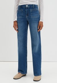 Jean en denim bleu avec une jambe droite, comportant des poches avant, une fermeture à bouton et une légère décoloration. Associé à des chaussons beige.