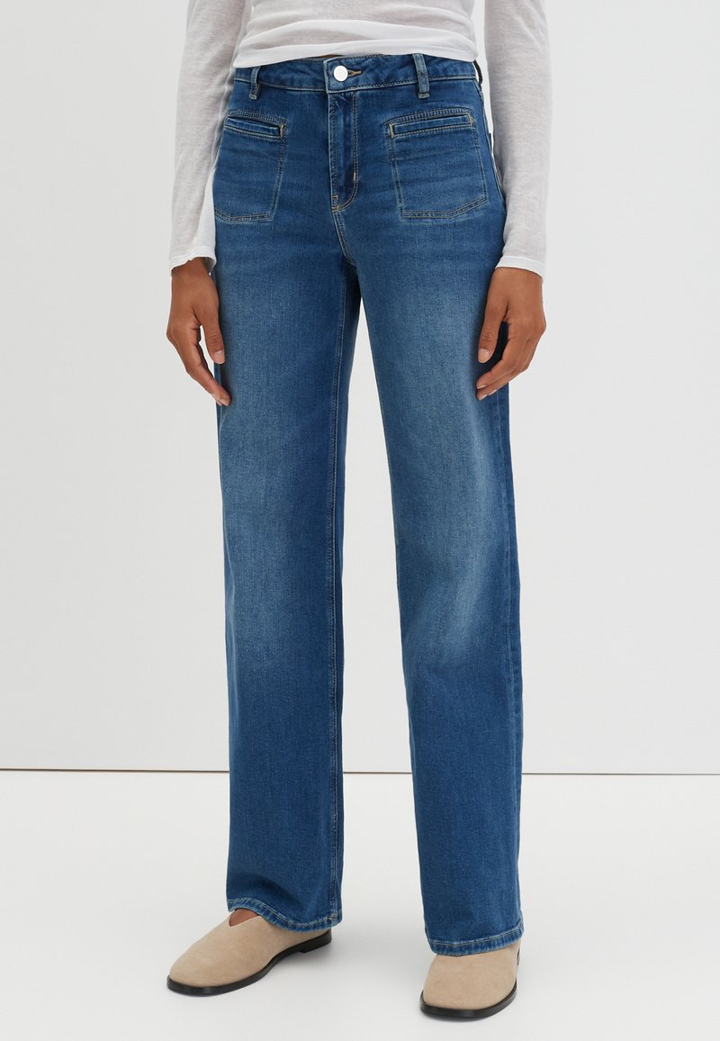 Jean en denim bleu avec une jambe droite, comportant des poches avant, une fermeture à bouton et une légère décoloration. Associé à des chaussons beige.