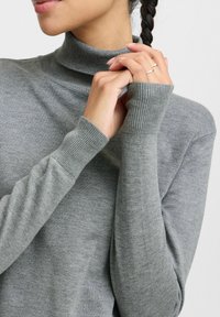 Pull à col roulé gris avec un col et des poignets côtelés, présentant une texture lisse et des manches ajustées. Les mains sont jointes devant.