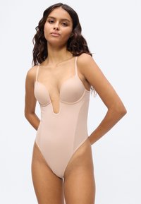 Γυναίκα που φοράει κορμάκι σε γυμνή απόχρωση με βαθύ V στο ντεκολτέ και λεπτές τιράντες στον ώμο, στέκεται μπροστά σε απλό ανοιχτόχρωμο φόντο.