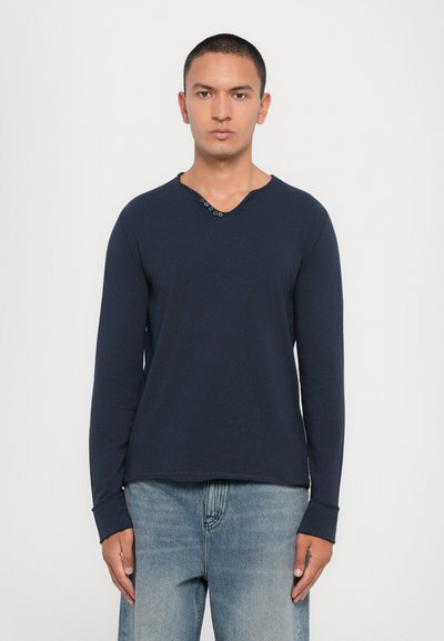 Navy long-sleeve T-shirt with a V-neck and three button accents, featuring a smooth texture and relaxed fit, paired with light denim jeans.