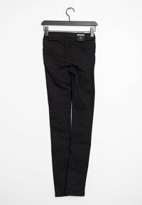 Jeans negros colgados en un gancho, con una textura suave, diseño de cinco bolsillos y un ajuste estrecho. Etiqueta visible en la parte trasera de la cinturilla.