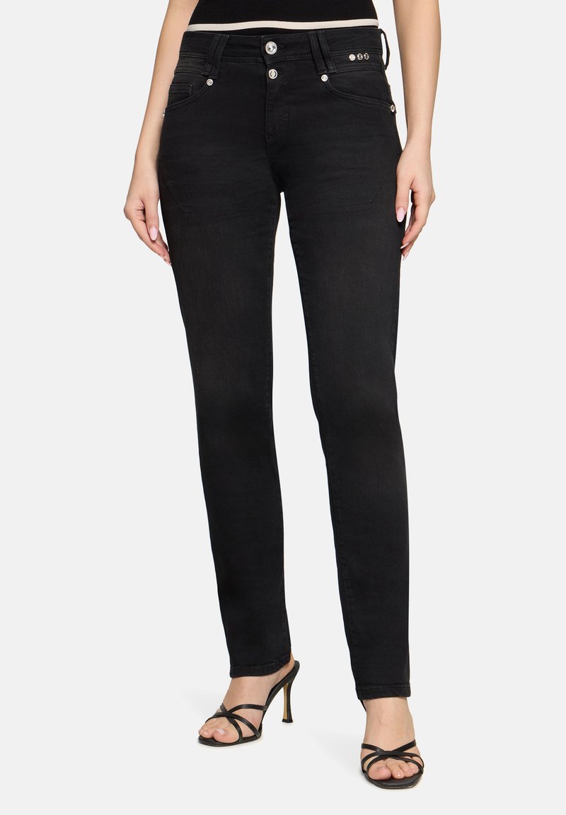 Femme portant un jean noir slim avec fermeture à double bouton et des sandales noires à talons hauts à brides, debout devant un fond blanc.