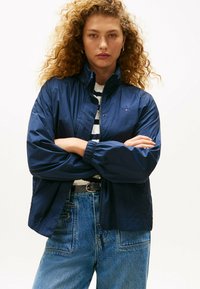 Granatowa, lekka kurtka z podniesionym kołnierzem i elastycznymi mankietami, w połączeniu z paskowanym swetrem noszonym pod spodem, zestawiona z niebieskimi dżinsami z wysokim stanem.