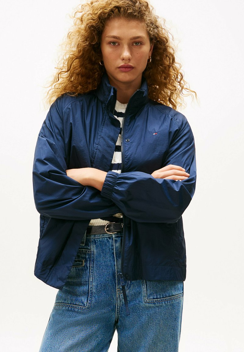 Granatowa, lekka kurtka z podniesionym kołnierzem i elastycznymi mankietami, w połączeniu z paskowanym swetrem noszonym pod spodem, zestawiona z niebieskimi dżinsami z wysokim stanem.