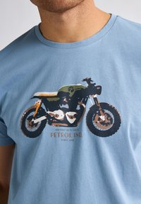 Světle modré bavlněné tričko s grafikou motocyklu v černé, hnědé a zelené barvě se textem "PETROL IND." ve spodní části.