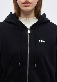 Κοντινό πλάνο ενός ατόμου που φοράει μαύρο φούτερ με φερμουάρ και τη λέξη "BOSS" στο στήθος, και από μέσα ένα λευκό ριγέ τοπ.