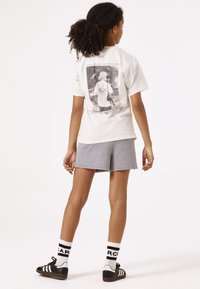 Camiseta de algodón blanca con un estampado gráfico en la parte posterior, que presenta un diseño de cerezas en tonos pastel, combinada con pantalones cortos grises y zapatillas negras.