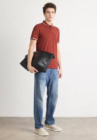 Červené polo tričko so prúžkovanými detailmi na golieri, v kombinácii so svetlomodrými džínsami. Model drží veľkú čiernu koženú tašku. Viditeľná je neformálna obuv.