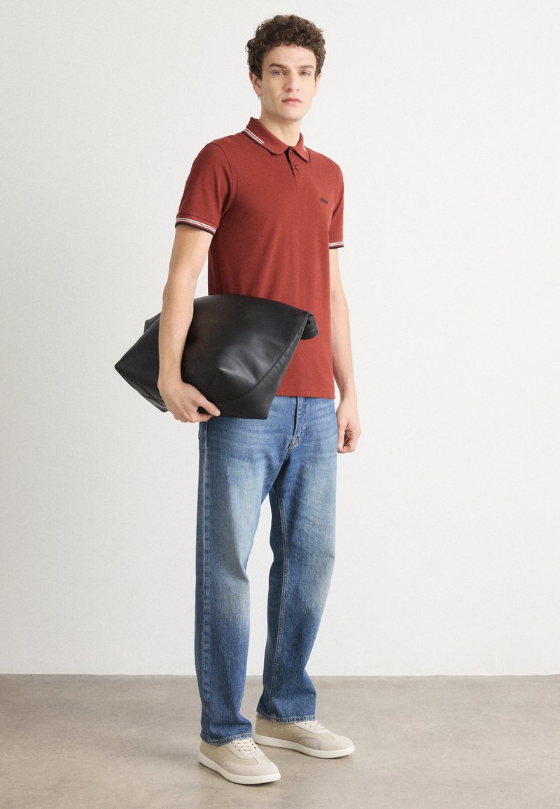 Polo rouge avec détails de col rayés, associé à un jean bleu clair. Le mannequin tient un grand sac en cuir noir. Chaussures décontractées visibles.