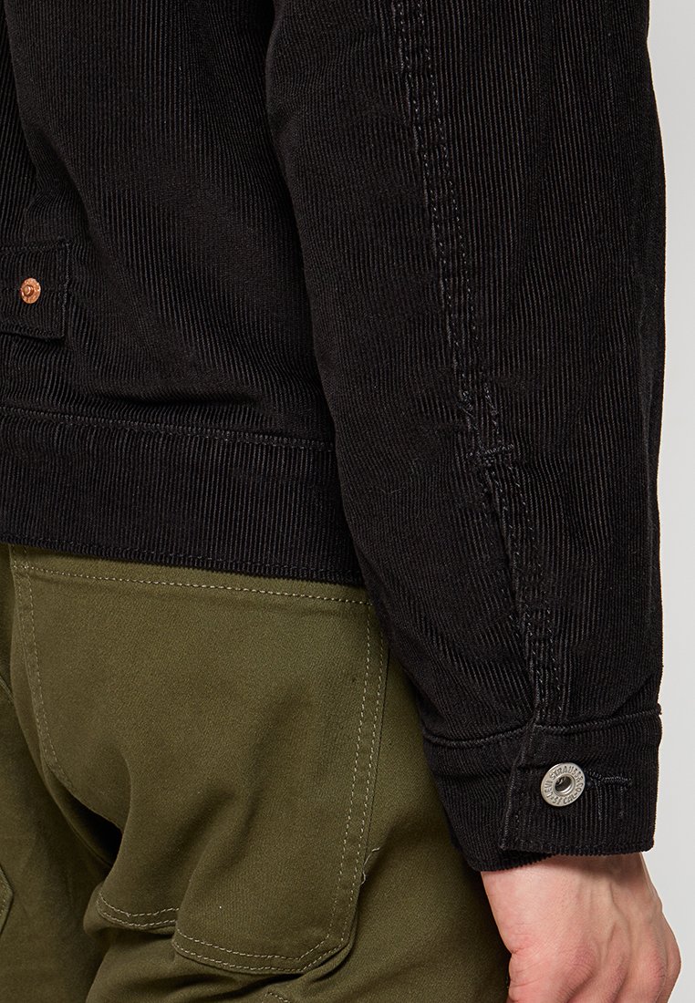 Chaqueta de pana negra con ribetes verticales, que presenta un puño con botón y pantalones de color verde oliva en contraste. Vista en primer plano de la textura de la tela.