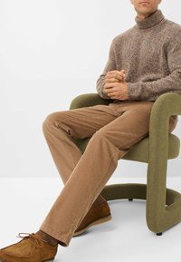Bruine corduroy broek gecombineerd met een gestructureerde bruine gebreide trui, zittend in een groene stoel met een ronde vorm, met tan schoenen.