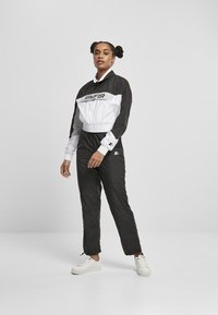 Veste coupe-vent noire et blanche avec un design court, dotée d'un col haut et de poignets élastiques, associée à un pantalon de survêtement noir assorti et des baskets blanches.