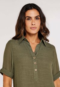 Olive green button-up shirt with a collared neckline, textured fabric, short rolled sleeves, and three wooden buttons on the front.