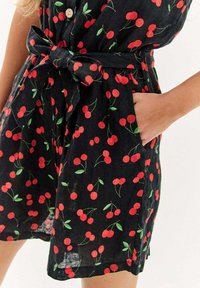 Černé šaty z látky s potiskem třešní v červené a zelené barvě, s páskem v pase a bočními kapsami, a předním zapínáním na knoflíky.