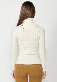 Cremfarbener Rippstrickpullover mit hohem, gefaltetem Kragen und Druckknöpfen. Taillierte Passform, lange Ärmel, kombiniert mit braunen Cordhosen.