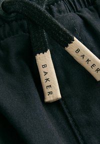 Čierna tkanina s elastickým pásom, obsahujúca textúrované čierne šnúrky a dve béžové štítky označené "BAKER."