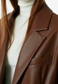 Brązowa marynarka z ekologicznej skóry z kołnierzem z wycięciem, pojedynczą kieszenią na piersi i gładką fakturą, zestawiona z białym prążkowanym golfem.