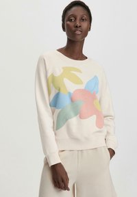 Hoodie vaaleanbeige, jossa on kukkakuva pinkillä, sinisellä, vihreällä ja keltaisella. Varusteena ribattuja hihansuita ja helmaa. Rentoa mitoitusta.