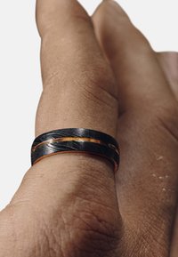 Svart og sølv sammenflettet ring med en kobberfargeaccent, vist på en hånd med en teksturert hudoverflate.