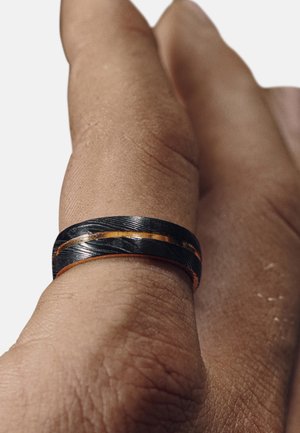 Svart og sølv sammenflettet ring med en kobberfargeaccent, vist på en hånd med en teksturert hudoverflate.