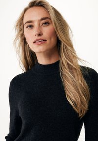 Černý pletený svetr s vysokým výstřihem, který má jemnou texturu a dlouhé rukávy, na osobě s dlouhými, světle hnědými vlasy.