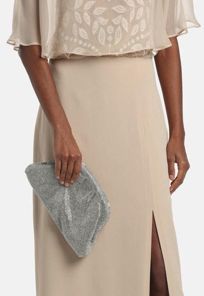 Pochette argentée métallique à surface texturée, tenue dans une main. Haut fluide beige avec un motif de feuilles brodé et jupe beige fendue.