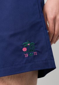 Navy shorts med grön och rosa broderad design som föreställer en växt, en rosa cirkel och texten "1972"; tillverkade av slätt tyg med en skräddarsydd passform.