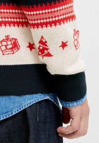Jersey de punto con un patrón en rojo y crema que presenta barcos, árboles y estrellas. Puños acanalados negros, superpuesto a una camisa de mezclilla, con jeans oscuros.