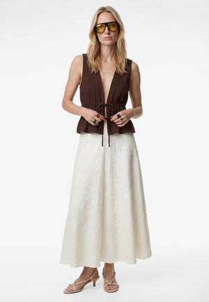 Vrouw met geel getinte zonnebril, mouwloze donkerbruine top met voorstrik, lange witte textuurrok en beige sandalen met hak.
