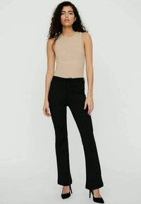 Top sin mangas beige combinado con pantalones acampanados negros de cintura alta y tacones negros, con un fondo gris claro.