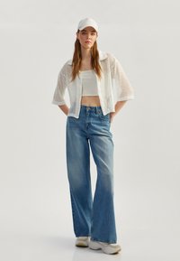 Chaqueta corta de punto abierto blanca sobre una blusa corta blanca, combinada con jeans de pierna ancha de talle alto azules y zapatillas de color claro.