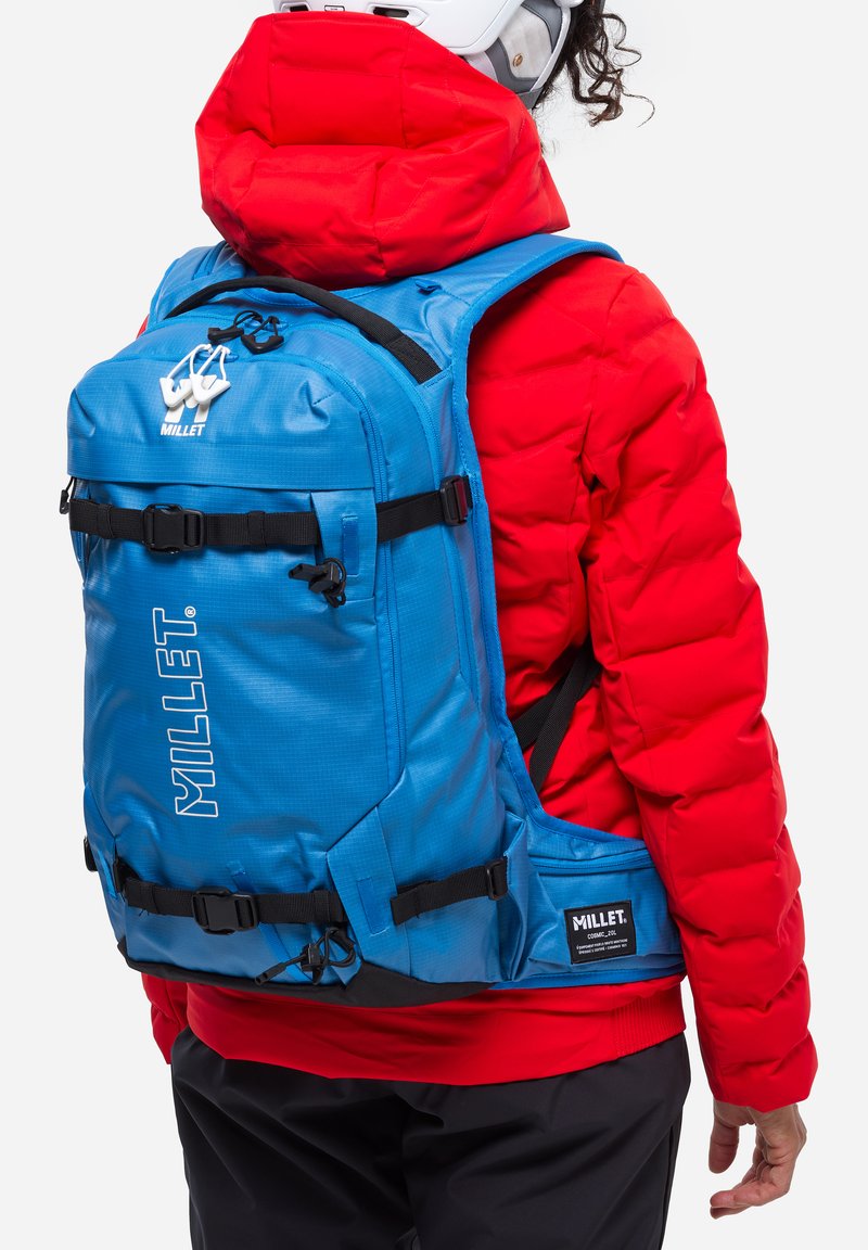 Le sac à dos de randonnée bleu dispose de bretelles noires ajustables, de plusieurs poches zippées et d'un logo blanc bien visible. Il est porté sur une doudoune rouge.