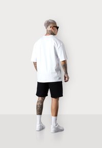 Weißes übergroßes T-Shirt mit kurzen Ärmeln, kombiniert mit schwarzen Shorts, weißen Turnschuhen und sichtbaren Tätowierungen an den Beinen. Zubehör beinhaltet Sonnenbrillen.