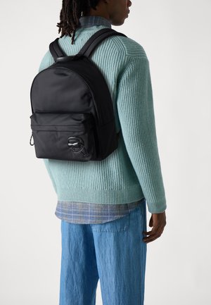 Black backpack made of smooth fabric, featuring a front pocket and minimalistic design. Paired with a textured light blue sweater and blue jeans.
