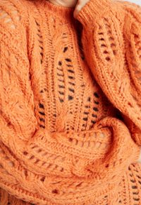 Pomarańczowy sweter dzianinowy z wzorem warkoczowym, z otwartymi splotami i teksturowaną powierzchnią. Miękki materiał o przytulnym, luźnym kroju.