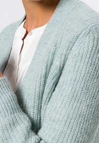 Světle modrý cardigan vyrobený z měkkého, texturovaného pleteného materiálu. Má dlouhé rukávy, hluboký výstřih do V a uvolněný střih.