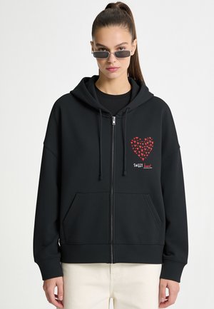 Femme portant de petites lunettes de soleil rectangulaires et un sweat à capuche zippé noir avec un cœur rouge composé de petits cœurs et le texte "SWEET heart".