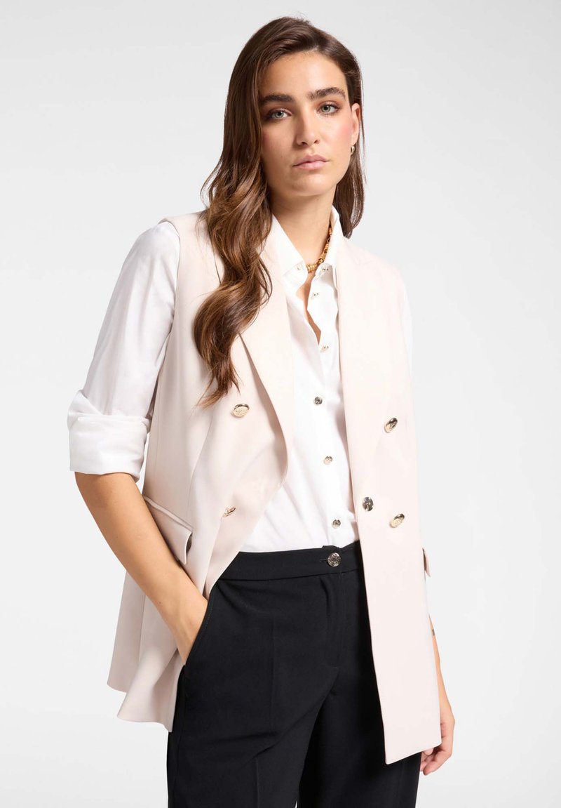 Helles pinker ärmelloses Blazer mit Doppelreiher-Design, goldenen Knöpfen und Taschen, getragen über einem weißen Hemd und schwarzen Hosen.