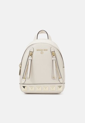 Cream leather backpack with two front zippers, gold hardware, and pyramid studs along the base. Rounded shape and top handle.