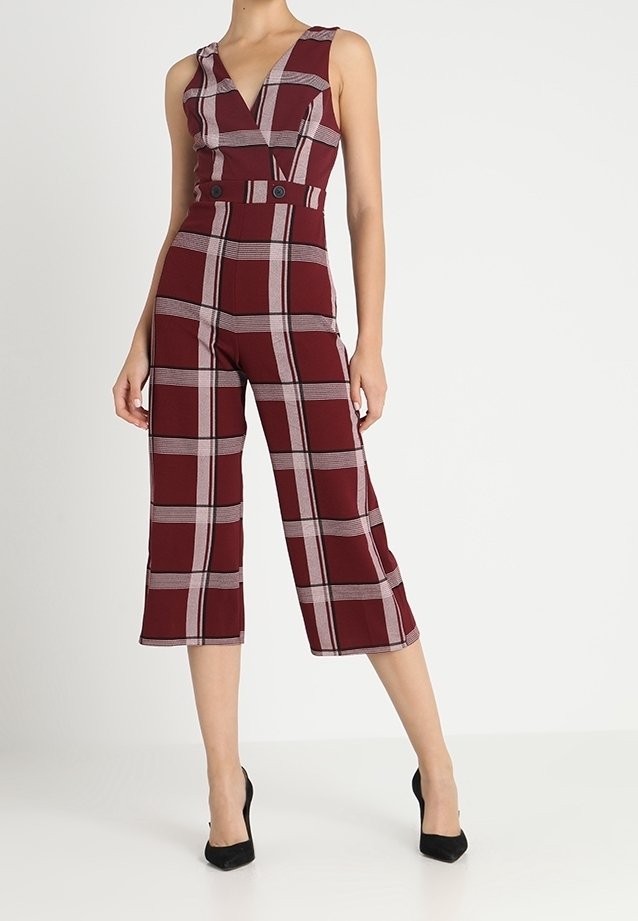 Combinaison bordeaux sans manches avec larges rayures à carreaux gris pâle, pantalon large raccourci, taille cintrée avec ceinture boutonnée, portée avec des talons noirs.