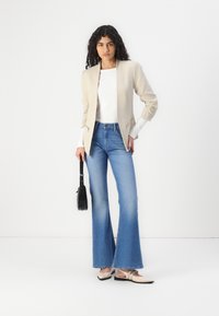 Blazer bej cu detalii pliate pe umeri, bluză albă cu mânecă lungă, blugi evazați de culoare albastră deschisă și pantofi bej cu talpă plată. Geantă de mână neagră cu curea.