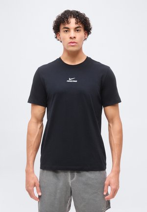 Jeune homme aux cheveux bouclés portant un t-shirt Nike Pro noir et un short gris, debout devant un fond clair uni.