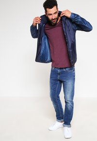 Marineblauer Windbreaker mit glänzender Oberfläche, kombiniert mit einem weinroten T-Shirt, dazu mittelblaue Jeans und weiße Sneakers.