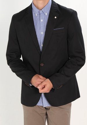 Mężczyzna w czarnym marynarce na niebieską, kraciastą koszulę zapinaną na guziki i beżowe spodnie, stojący z dłońmi złączonymi z przodu.