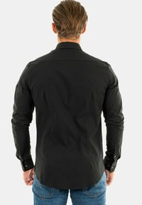 Mężczyzna z jasnobrązowymi włosami, ubrany w czarną koszulę z długim rękawem i niebieskie dżinsy, stojący tyłem na tle gładkiego białego tła.